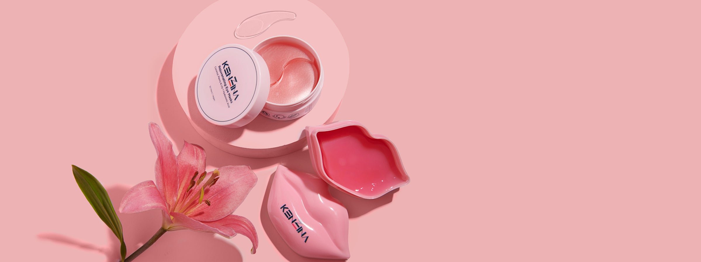 Two pink open jars of Kenzina products, eye masks and lip masks with a flower on a pink background